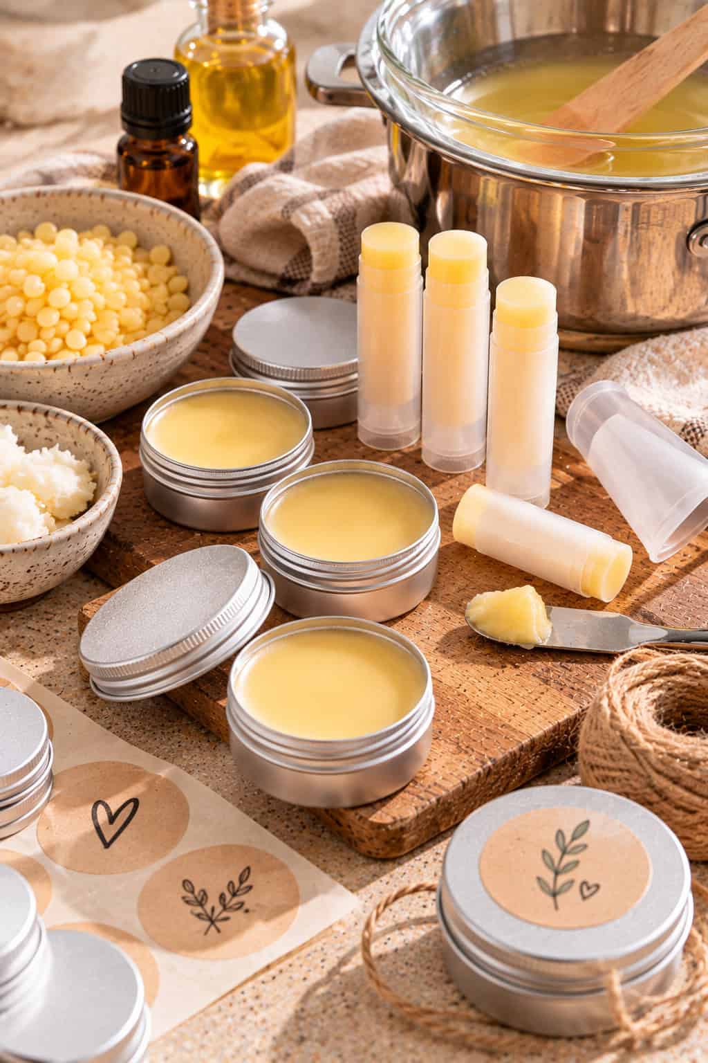 Selbstgemachter Lippenbalsam wird in kleine Döschen und Pflegestifte abgefüllt, daneben liegen Bienenwachs, Sheabutter, Öl und runde Aufkleber zum Beschriften.