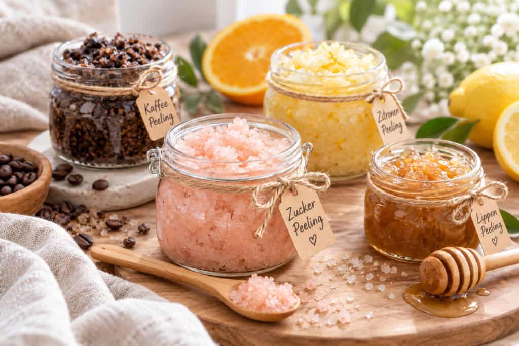 Mehrere selbstgemachte Peelings in kleinen Gläsern auf einem Holztisch, darunter Kaffeepeeling, Zuckerpeeling und fruchtige Varianten mit Zitrone und Orange. Daneben liegen Kaffeebohnen, Honig, ein Holzlöffel und helle Stoffe.